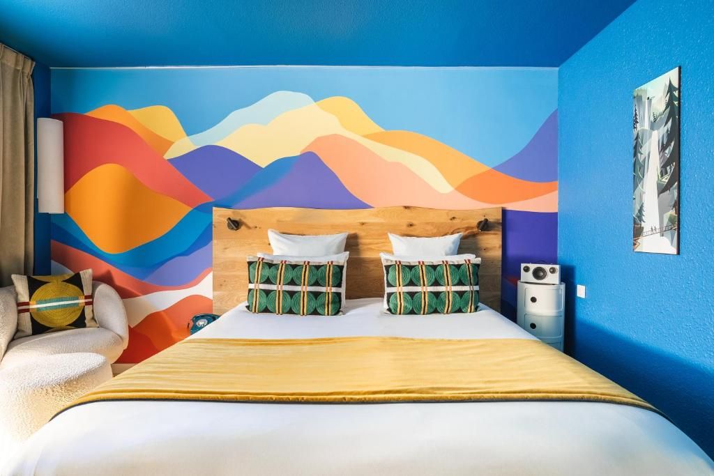 Une chambre dont le mur porte une fresque de montagnes en aplats de couleur, avec son grand lit et son bureau