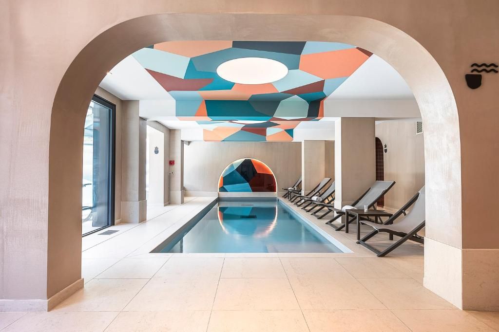 La piscine intérieure sous sa voûte blanche et son plafond à motifs colorés, bordée de transats