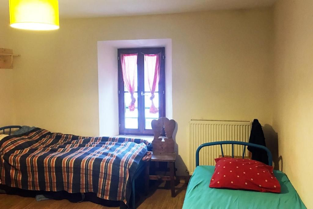 Une chambre simple aux murs clairs, avec ses lits, sa fenêtre à rideaux et son radiateur